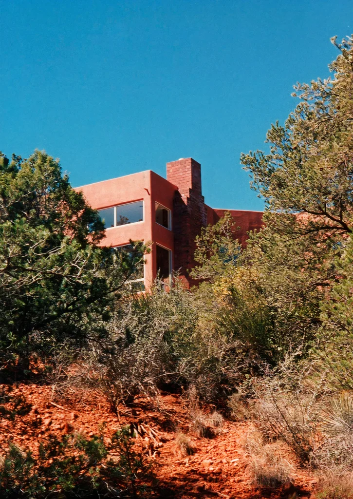 Two-story red stucco house emerging above dense juniper and desert shrubs on a steep red-rock hillside under a bright blue sky.