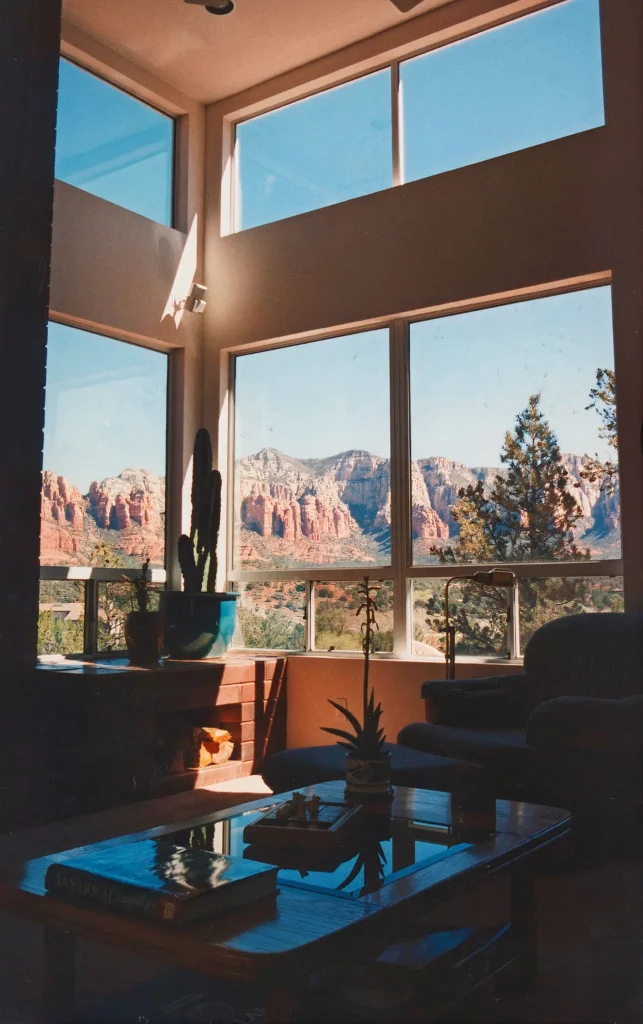Double-height living room with tall corner windows framing dramatic red rock cliffs, cactus in a blue pot, and seating arranged around a glass coffee table.