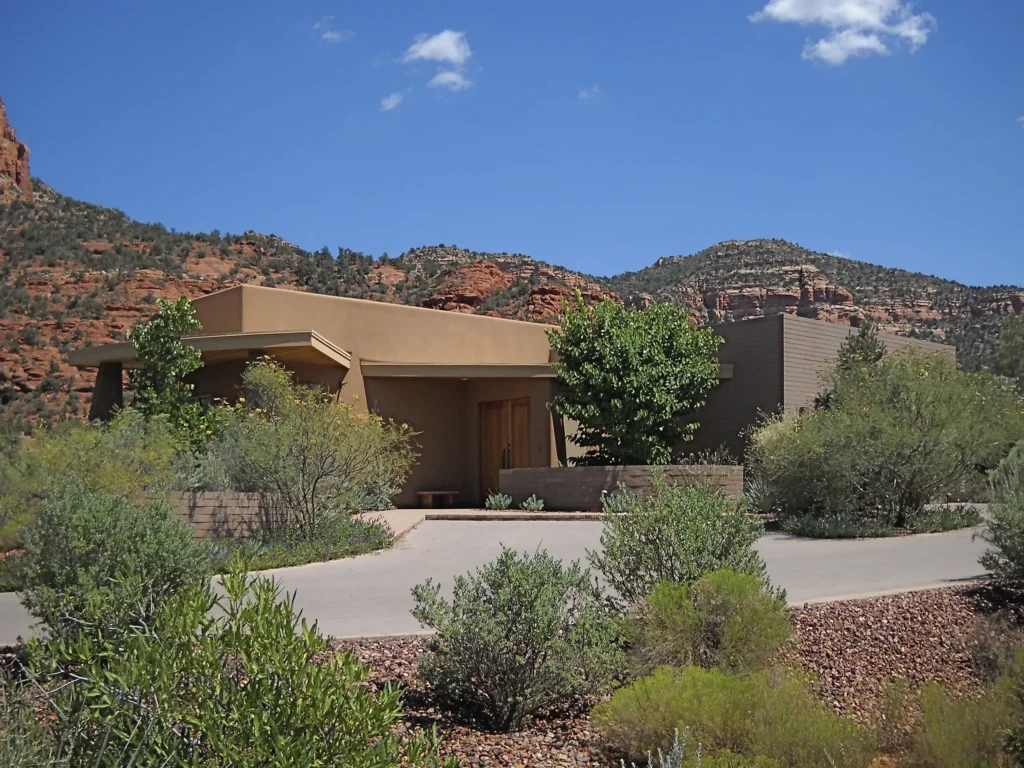 Modern stucco residence nestled in desert vegetation with a paved drive and dramatic red rock cliffs in the background.