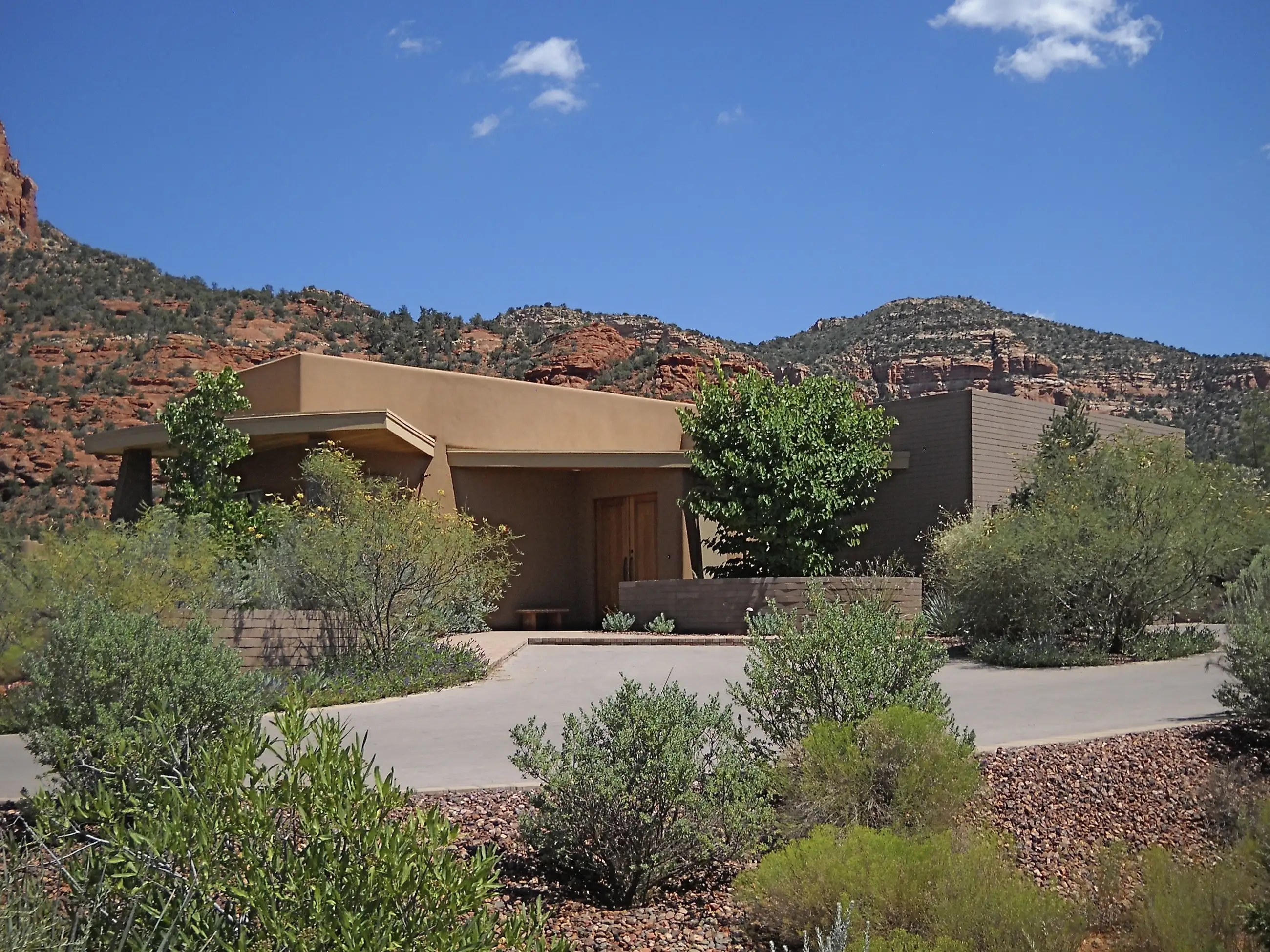 Modern stucco residence nestled in desert vegetation with a paved drive and dramatic red rock cliffs in the background.