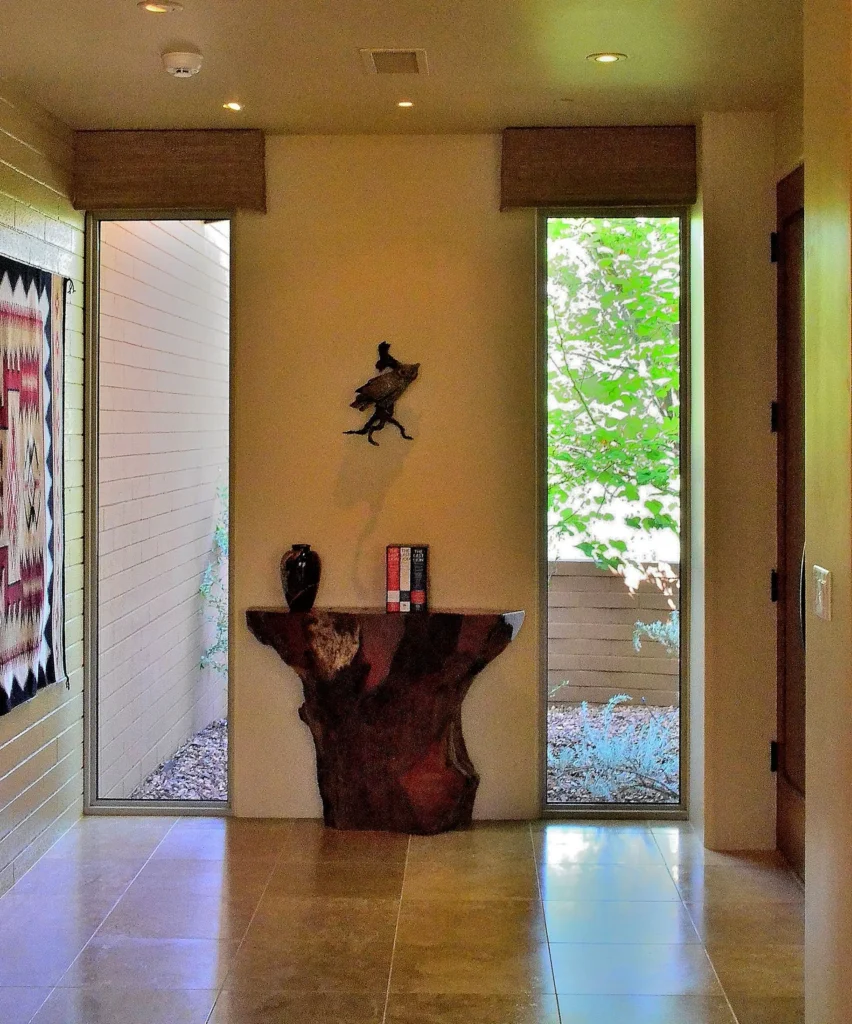Warm entry hall with stone tile floor, sculptural wood console table, tall narrow windows framing greenery, and a Navajo-style textile hanging on the wall.