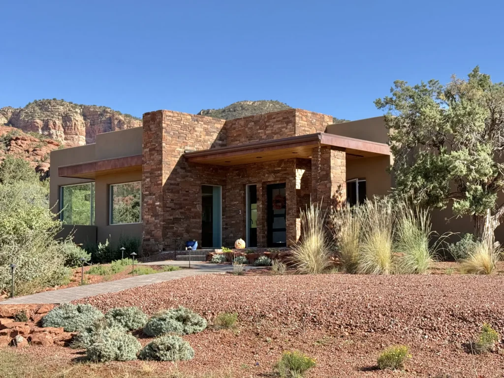 Angled view of low-profile stucco and stone house with large windows and native plantings in the foreground.