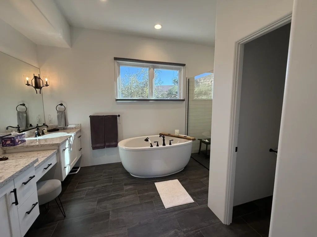 Spacious primary bathroom with freestanding soaking tub beneath a window overlooking desert trees and hills.
