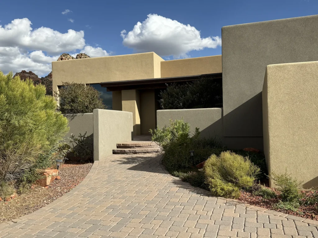 Curved paver walkway leading to a modern stucco entry courtyard framed by desert landscaping and big sky clouds.