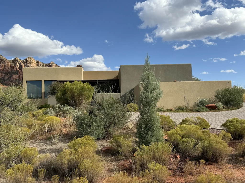 Modern stucco residence nestled in natural high-desert vegetation with red rocks behind.