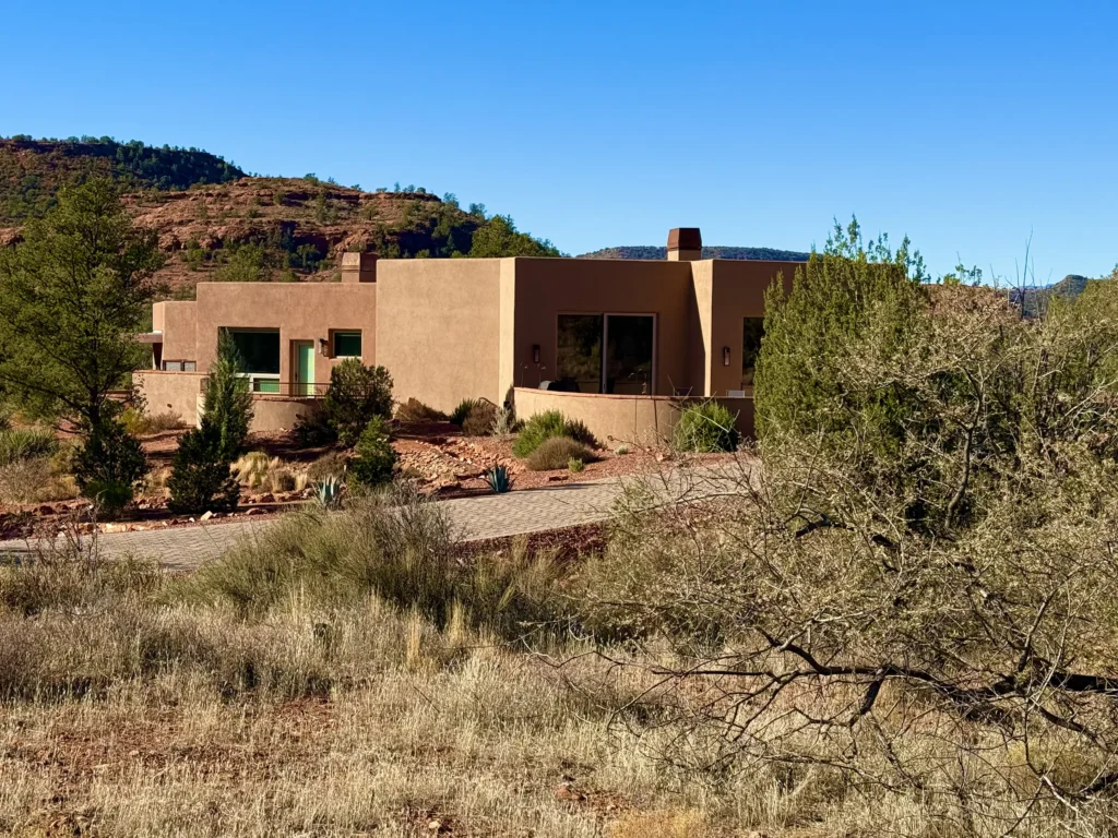 Stucco home viewed through desert foliage with red rock terrain in the background.